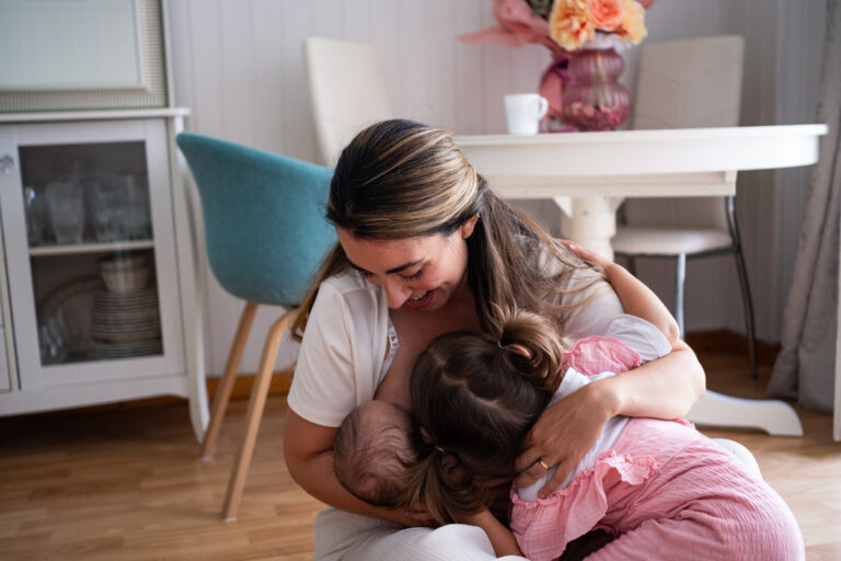 A mother sits on the floor in a warm living room, breastfeeding her baby while her toddler hugs her closely. The environment is bright and inviting, accented by soft colors and decor.